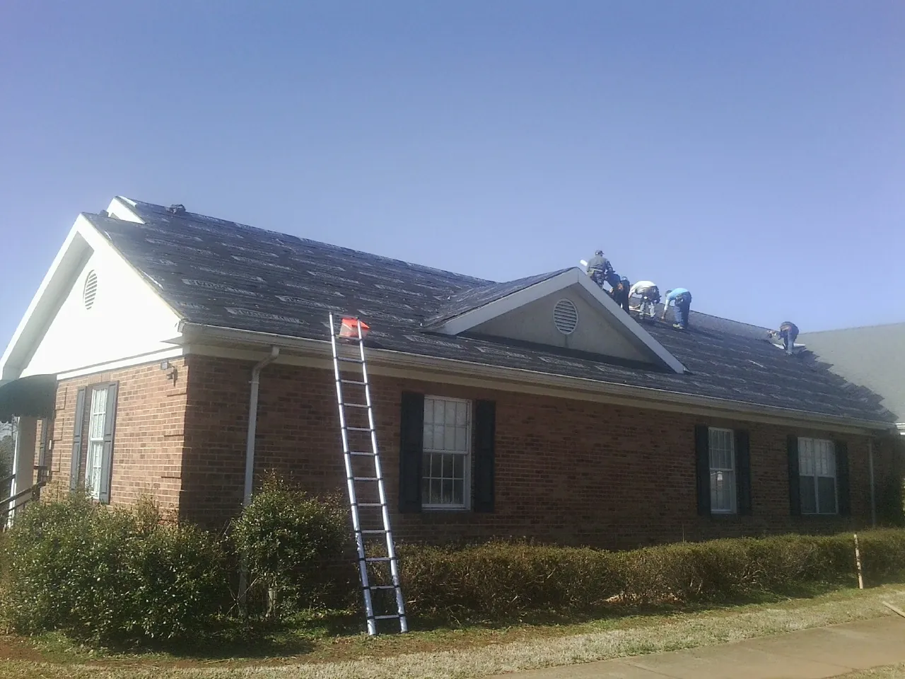 Crew tearing shingles off a brick ranch roof with a ladder set — Metal Roof Repair work by our crew in Cove City