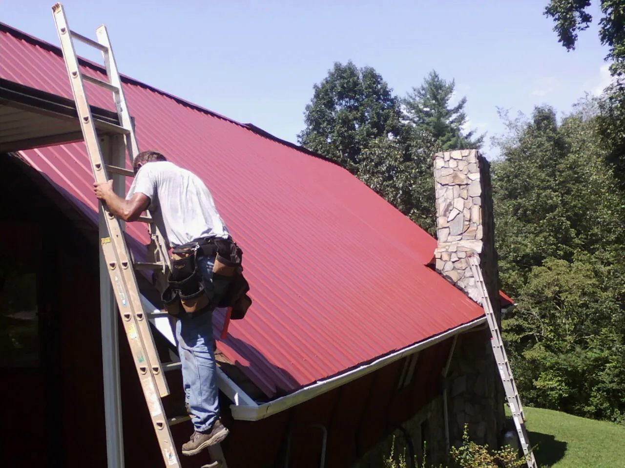 Roofer on a ladder beside a red metal roof and stone chimney — Soffit & Fascia Repair by Roofing Brothers in Cove City